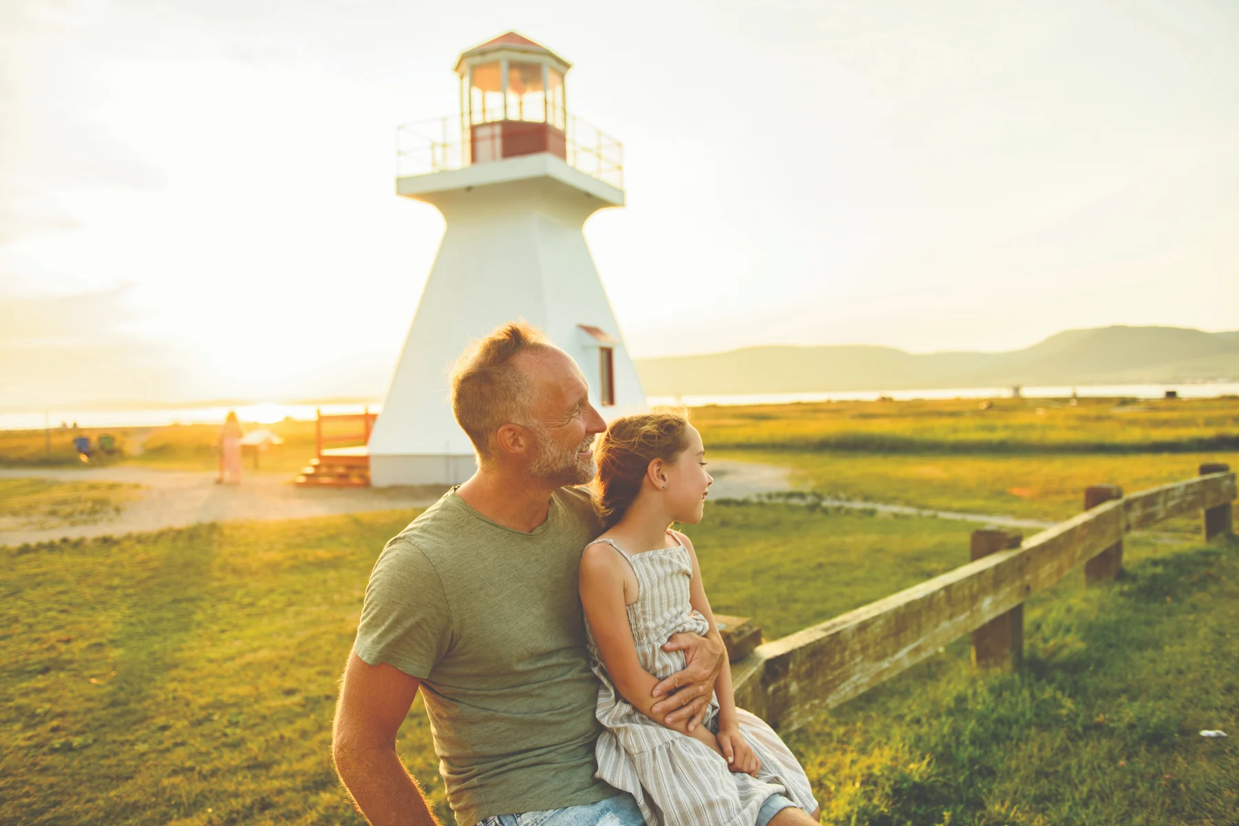 Un père et sa fille devant un phare, dans la lumière du soleil couchant.